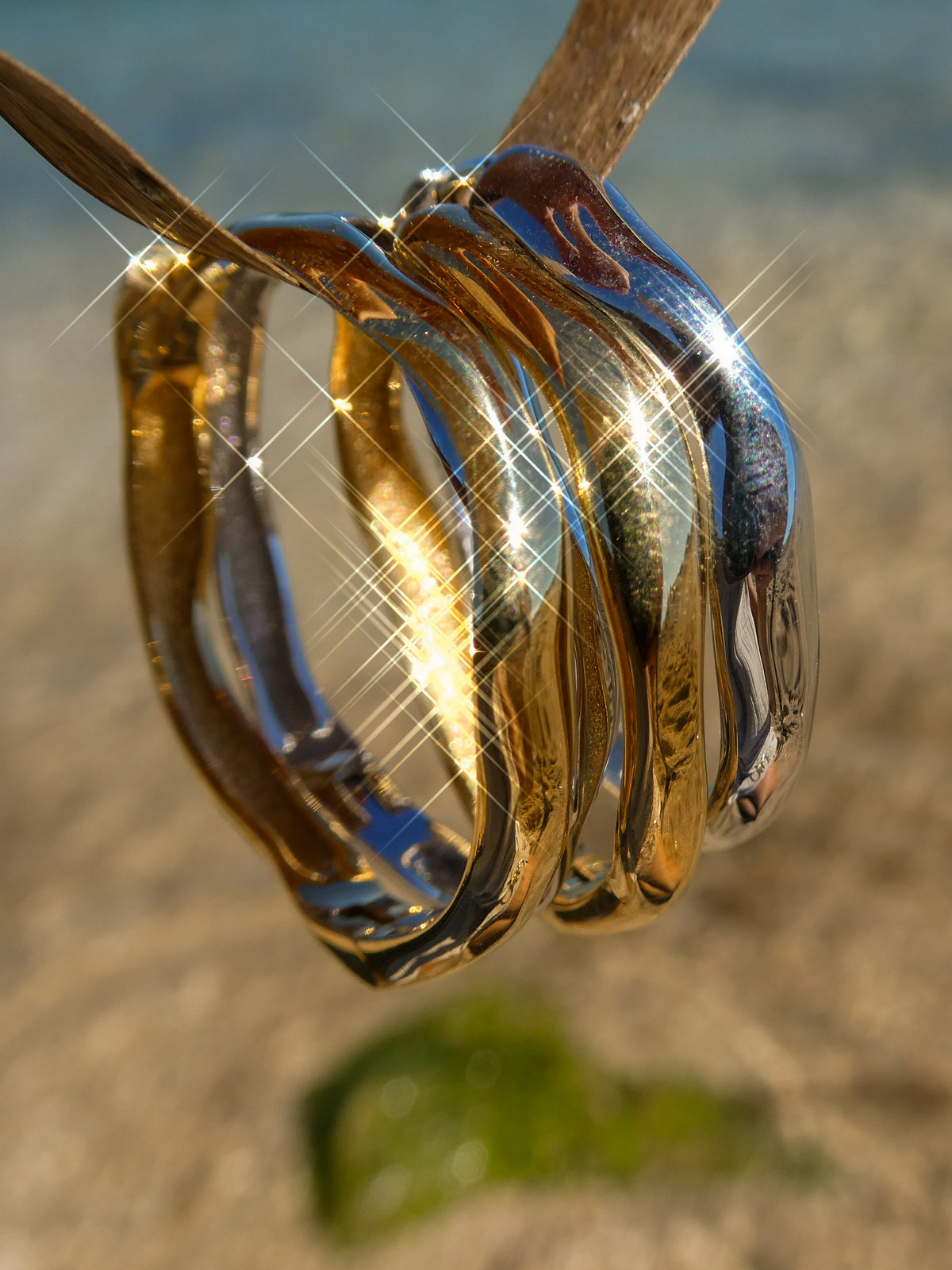A close-up of three reflective metallic rings hanging from a wooden branch outdoors, with a blurred sandy and water background and bright starburst light effects.