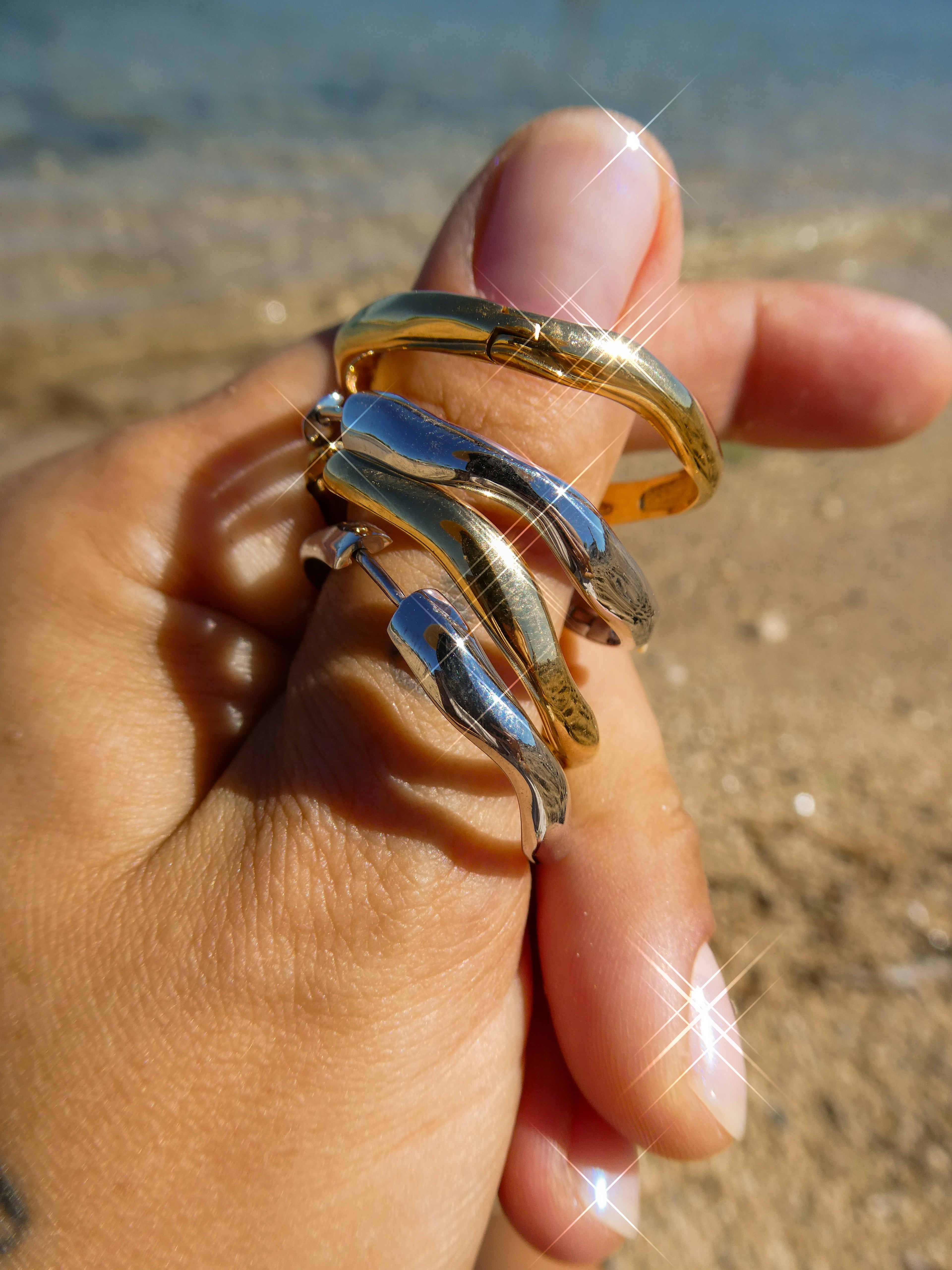 Nesoí hoops in gold and silver with blue accents, stacked on finger with Mediterranean Sea and beach in background