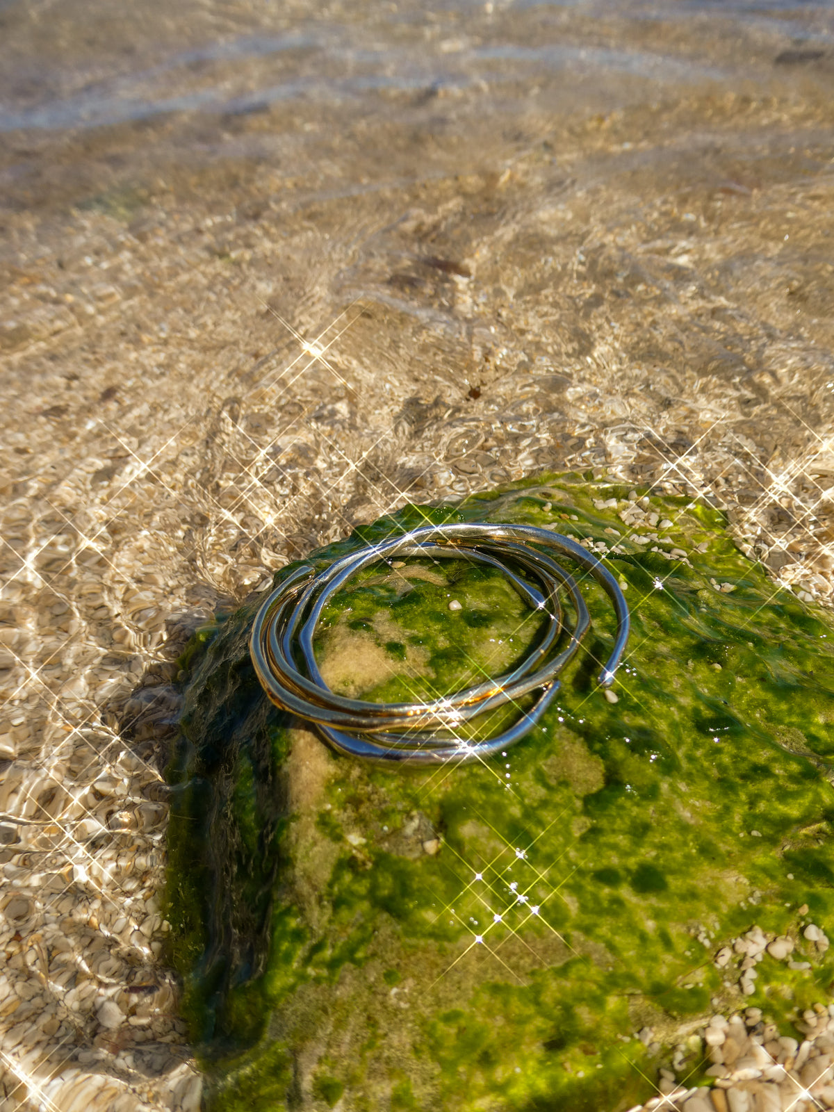 Nyx bracelets in gold and silver with blue accents, stacked on moss-covered stone on a Mediterranean sea beach